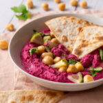 Close-up of Beetroot hummus decorated with chickpeas and pita bread on pink towel on the white wooden table